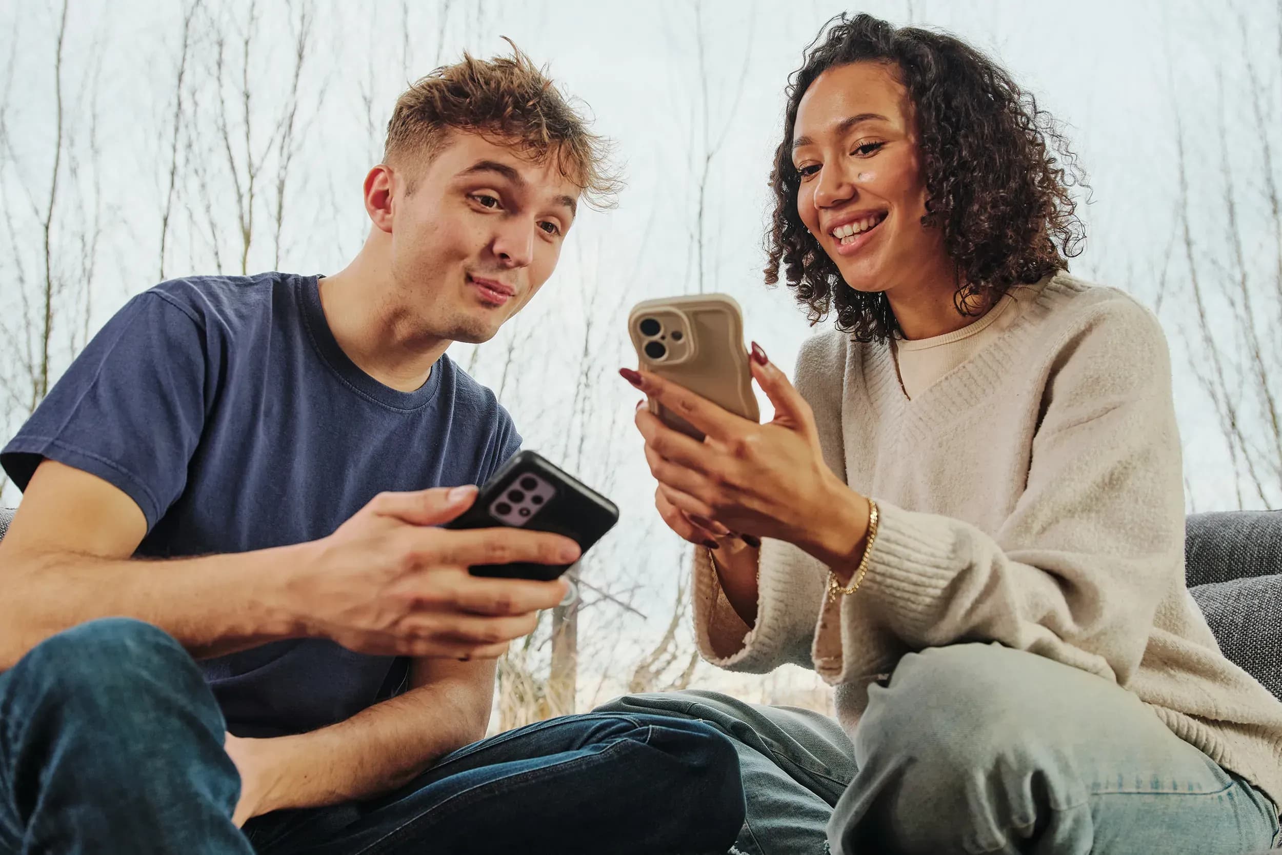 Smiling young man and woman sitting together, enthusiastically looking at each other’s smartphones.