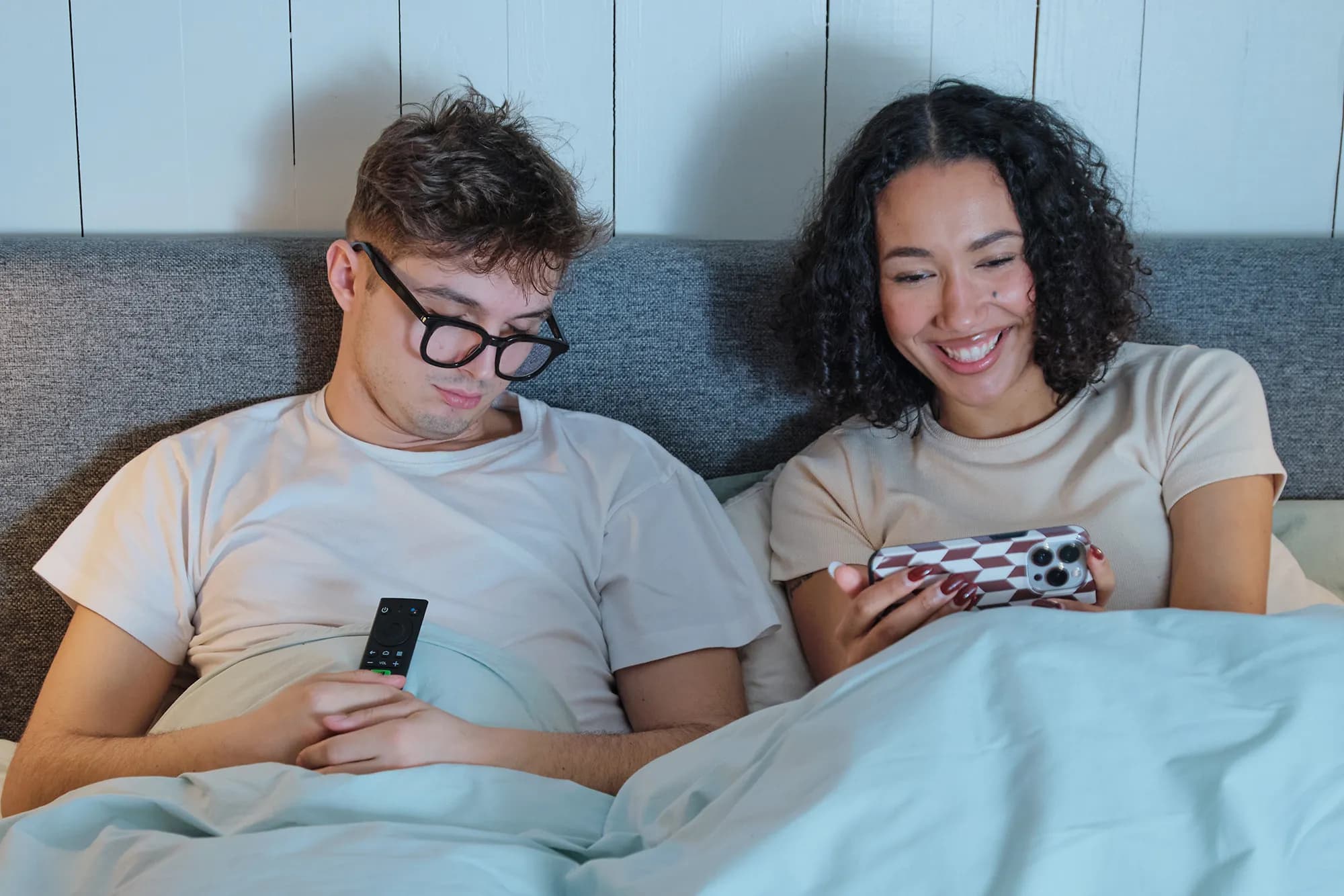An image of a man asleep with TV remote in hands and a woman next to him watching TADAAM TV on a phone